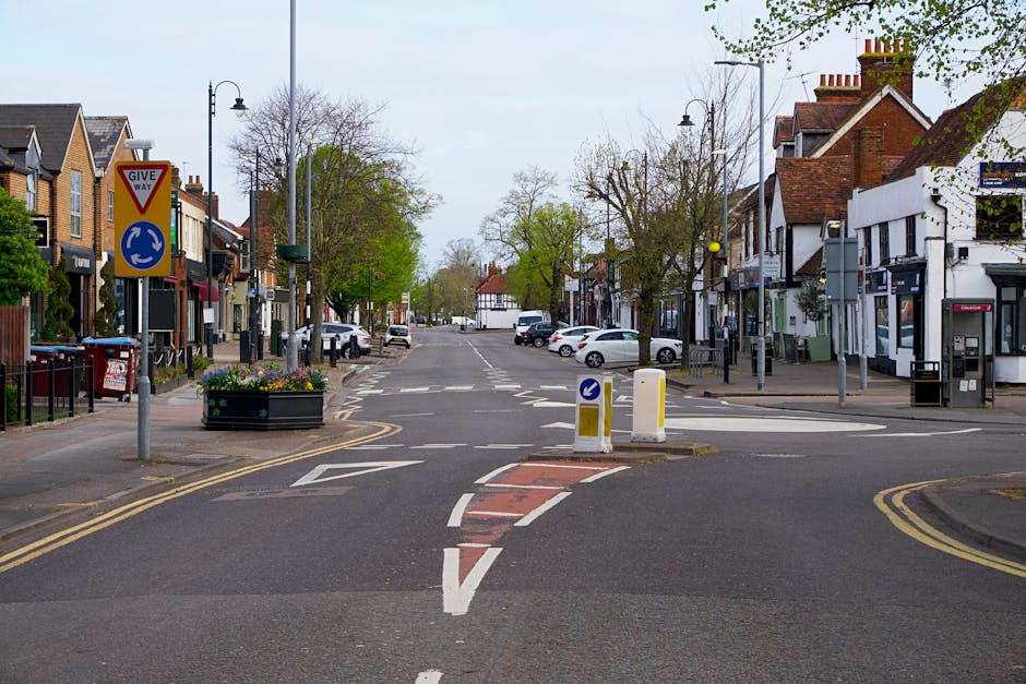 A quiet street scene in Kings Langley High Street, featuring a central crossing with white and red markings, surrounded by small shops and residential buildings with pitched roofs. The street is lined