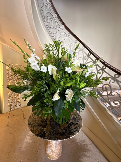 A tall, lush floral arrangement in a round, black vase placed on a dark marble-topped table, featuring a mix of white flowers including lilies and anthuriums, complemented by various green foliage and
