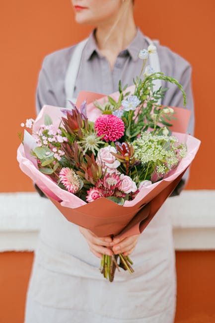 A person wearing a light grey shirt and white apron is holding a large bouquet of fresh flowers wrapped in pink and terracotta paper, with a mix of pink, white, and green blooms including chrysanthemu