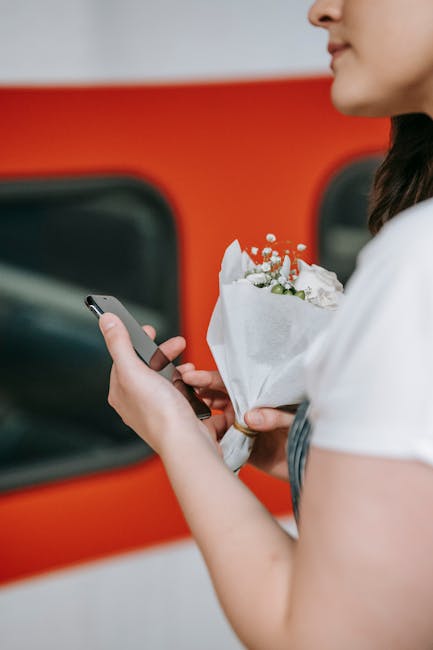 A woman holding a bouquet of fresh white and pastel pink flowers wrapped in white paper, while using her smartphone. The bouquet includes roses, baby's breath, and other delicate blooms, with green fo