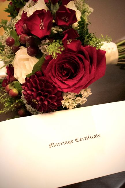 A close-up view of a floral arrangement featuring a large, deep red rose surrounded by smaller white and red flowers, including dahlias and hypericum berries, with fresh green foliage. The bouquet is 
